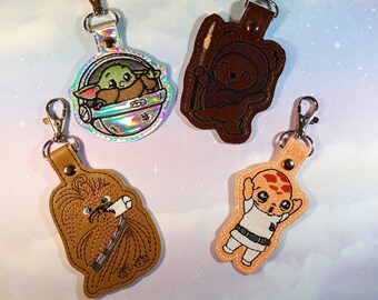 May include: Four keychains featuring characters from Star Wars. The keychains are made of brown fabric and have embroidered details. The characters include a baby Yoda in a pod, a furry creature, a character with a brown hat, and a character with a white shirt.