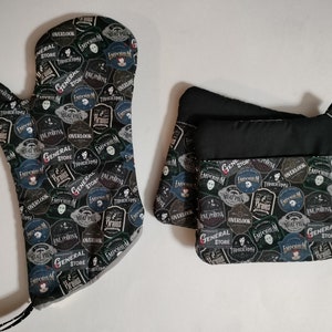 May include: A set of two black oven mitts with a pattern of black and white labels. The labels have text that reads "General Store", "Emporium", "Overlook", "Meat Pies", "Frains", and "Tridermy".