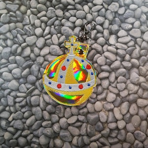 Może przedstawiać: Złoty i tęczowy brelok do kluczy z motywem korony. Korona ma czerwone i niebieskie akcenty i jest otoczona srebrną wstążką.