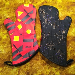 Puede incluir: Dos guantes para horno, uno rojo con formas geométricas negras y detalles amarillos, y otro negro con detalles morados y dorados.