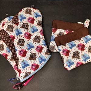 Puede incluir: Un juego de textiles de cocina con un diseño caprichoso de búhos. El juego incluye un guante de horno y dos agarraderas. Los artículos son blancos con un patrón repetido de búhos en varios colores y lunares. El guante de horno tiene un acento de lunares marrones.