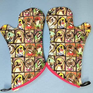 May include: Pair of oven mitts with a colorful stained-glass design featuring anime characters. The mitts have a black background with a repeating pattern of figures in various poses. The edges are trimmed with bright pink fabric.
