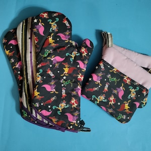 May include: Two oven mitts with a black background and a colorful cartoon pattern. One mitt is a double layer with a purple and white striped fabric on the outside. The other mitt is a single layer with a pink lining.