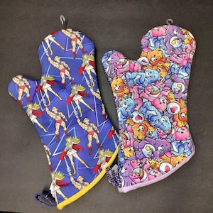 May include: Two oven mitts. One is blue with a repeating pattern of a muscular man and a woman with a sword. The other is covered in a colorful pattern of cartoon bears and other characters. Both have hanging loops.