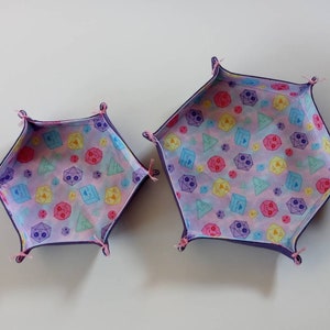 May include: Two purple fabric dice trays with a pink, blue, and yellow dice pattern. The smaller tray is on the left and the larger tray is on the right.