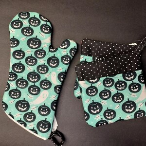 May include: A teal oven mitt and pot holder set with a Halloween theme. The set features a pattern of black jack-o'-lanterns and white ghosts on a teal background. The pot holder has a black and white polka dot pattern on the top.