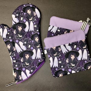 May include: A set of two purple oven mitts with a black and white print of a witch with a camera and ghosts. The mitts are made of fabric and have a quilted texture.