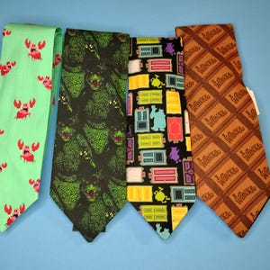 May include: Four different patterned neckties. The first tie is green with pink crabs. The second tie is black with green monster faces. The third tie is black with colorful doors and a monster. The fourth tie is brown with a chocolate bar pattern that says "Wonka".