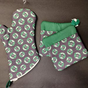 Peut inclure: Un ensemble de trois maniques de four vertes et noires avec un motif répété d'un cercle avec une croix à l'intérieur. Les maniques sont en tissu et ont une bordure blanche.