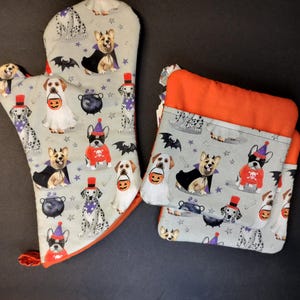 May include: Set of two Halloween-themed oven mitts and pot holders. The fabric features a light gray background with a pattern of dogs dressed in costumes. The dogs are wearing hats, capes, and holding candy buckets. The mitts and pot holders have orange trim.