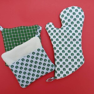 May include: Three green and white oven mitts. One mitt is a potholder with a plaid pattern. Another potholder has a white background with a green pattern of a coffee cup logo. The third mitt is a single oven mitt with a white background and a green pattern of a coffee cup logo.