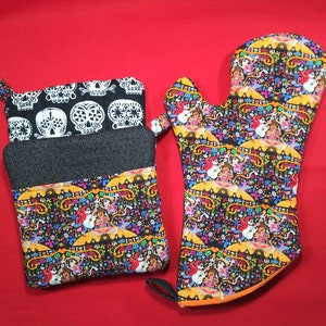 Puede incluir: Un juego de dos manoplas de horno negras y blancas con un patrón colorido del Día de Muertos. Las manoplas están hechas de tela y tienen un lazo para colgar.