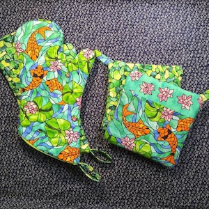 Puede incluir: Dos agarraderas de olla coloridas con un diseño de vitral que presenta peces naranjas y hojas verdes. Las agarraderas de olla están hechas de tela y tienen un lazo para colgar.