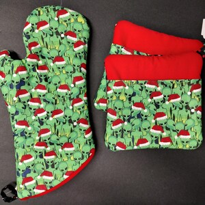 May include: A set of two green and red oven mitts with a pattern of aliens wearing Santa hats. The mitts are made of fabric and have a red trim. The smaller mitt is a potholder. The tag says "$14".