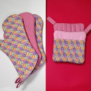 May include: A set of colorful oven mitts and a pot holder. The items feature a repeating pattern of heart-shaped designs with the word "BUTTS" in a playful font. The mitts and pot holder have pink accents.