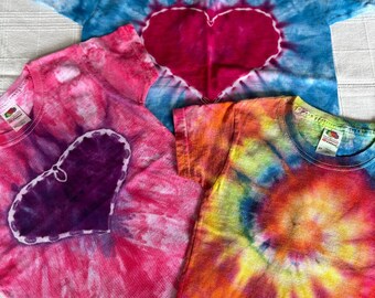 May include: Three tie-dye t-shirts with a heart design in the center of each shirt. The shirts are in shades of blue, pink, and orange.