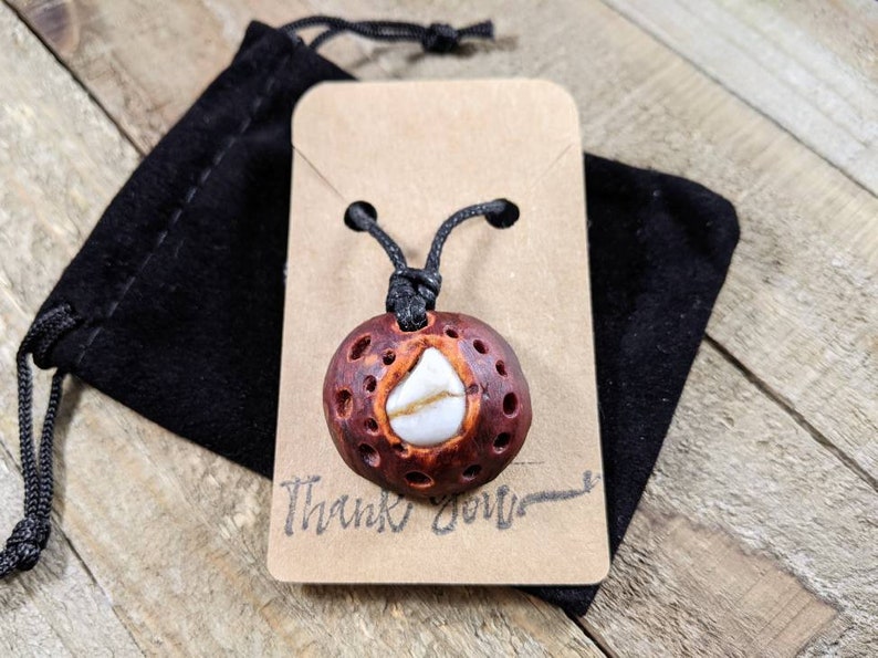 May include: A brown and white pendant with a black cord, displayed on a brown card with the text "Thank You". The pendant is made of wood and has a white stone in the center.