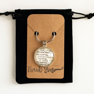 May include: A silver keychain with a glass charm featuring a dictionary definition of the word "caustic". The keychain is displayed on a brown card with the words "Thank You" written in black ink.