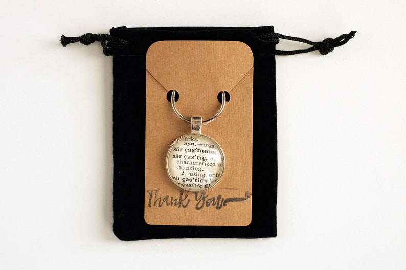 May include: A silver keychain with a glass charm featuring a dictionary definition of the word "caustic". The keychain is displayed on a brown card with the words "Thank You" written in black ink.