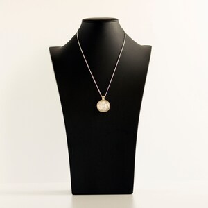 May include: A silver chain necklace with a round, gold pendant on a black mannequin bust. The pendant is covered in small, white stones.