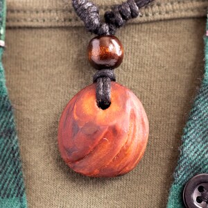 May include: A close-up of a wooden pendant necklace. The pendant is a reddish-brown color with a carved design and a hole in the center. The necklace has a black cord and a wooden bead. The background includes a green and black plaid shirt.