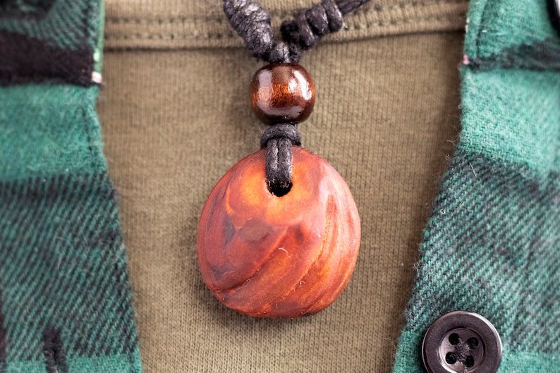 May include: A close-up of a wooden pendant necklace. The pendant is a reddish-brown color with a carved design and a hole in the center. The necklace has a black cord and a wooden bead. The background includes a green and black plaid shirt.