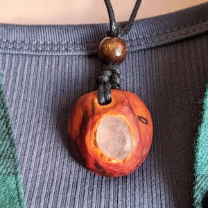 Pu&ograve; includere: Collana con pendente in legno fatto a mano. Il pendente marrone rossiccio intagliato ha una cavit&agrave; centrale con una pietra chiara. La collana ha un cordino nero e una perla di legno. Il pendente misura circa 3 cm di diametro.