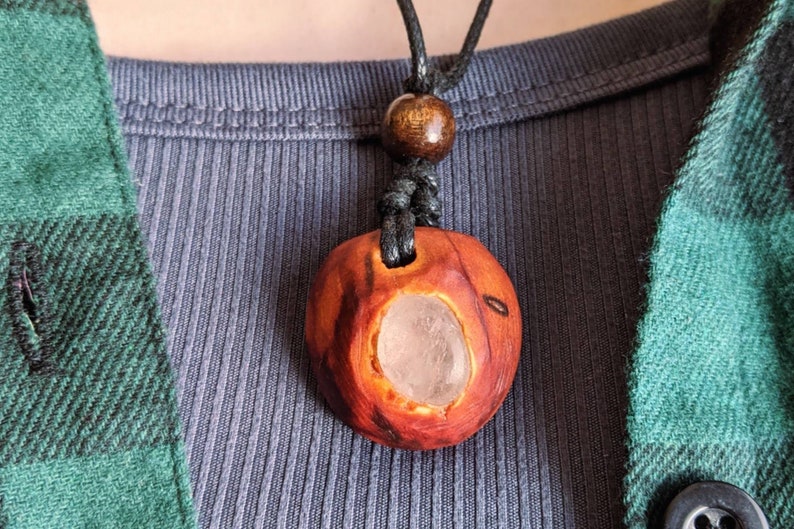 Pu&ograve; includere: Collana con pendente in legno fatto a mano. Il pendente marrone rossiccio intagliato ha una cavit&agrave; centrale con una pietra chiara. La collana ha un cordino nero e una perla di legno. Il pendente misura circa 3 cm di diametro.