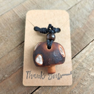 May include: A brown and white mushroom-shaped pendant necklace with a black cord and a brown wooden bead. The pendant has a white glaze and a brown base. The necklace is displayed on a brown card with the text "Thank You" written on it.
