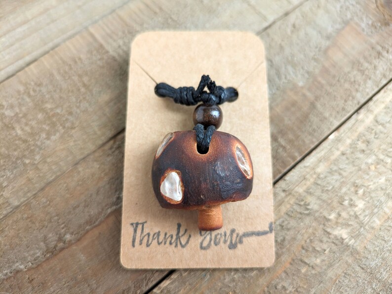 May include: A brown and white mushroom-shaped pendant necklace with a black cord and a brown wooden bead. The pendant has a white glaze and a brown base. The necklace is displayed on a brown card with the text "Thank You" written on it.
