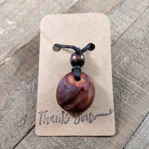 May include: A handcrafted wooden pendant necklace with a dark brown, oval-shaped pendant. The pendant is suspended from a black cord with a wooden bead accent. The necklace is displayed on a small, brown card with the words "Thank You".