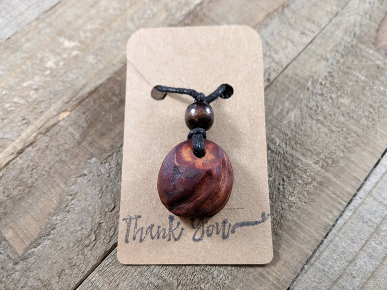 May include: A handcrafted wooden pendant necklace with a dark brown, oval-shaped pendant. The pendant is suspended from a black cord with a wooden bead accent. The necklace is displayed on a small, brown card with the words "Thank You".