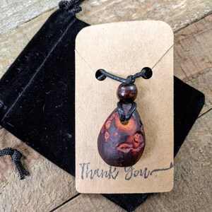 May include: A wooden pendant necklace with a brown and red teardrop-shaped pendant. The pendant has a carved design and is on a black cord with a brown wooden bead. The necklace is displayed on a brown card with the text "Thank You".