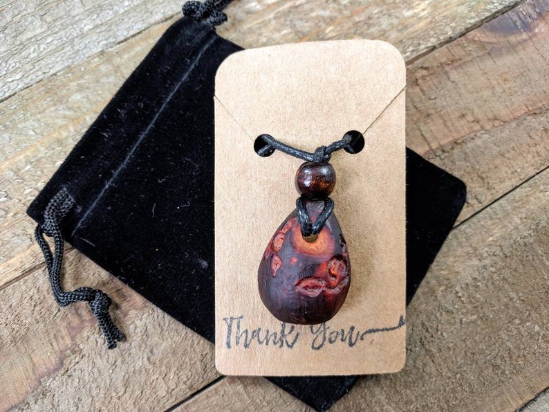 May include: A wooden pendant necklace with a brown and red teardrop-shaped pendant. The pendant has a carved design and is on a black cord with a brown wooden bead. The necklace is displayed on a brown card with the text "Thank You".