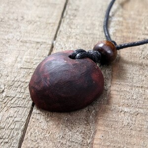 May include: A handcrafted necklace featuring a dark red, oval-shaped pendant with a hole. The pendant is suspended from a black cord, with a wooden bead accent. The necklace rests on a rustic wooden surface.