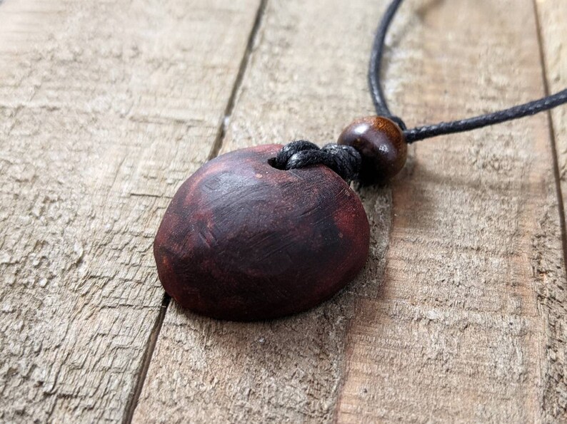 May include: A handcrafted necklace featuring a dark red, oval-shaped pendant with a hole. The pendant is suspended from a black cord, with a wooden bead accent. The necklace rests on a rustic wooden surface.
