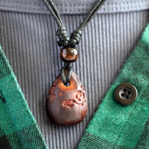May include: A dark brown wooden pendant with intricate carvings hangs from a black cord necklace. The necklace is worn over a gray shirt and a green plaid shirt.