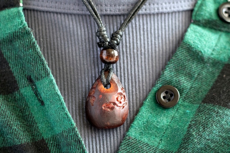 May include: A dark brown wooden pendant with intricate carvings hangs from a black cord necklace. The necklace is worn over a gray shirt and a green plaid shirt.