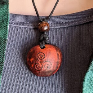 May include: A round, reddish-brown pendant necklace with a black cord. The pendant features a triple spiral design in black. The necklace is worn over a gray ribbed shirt and a green and black plaid shirt.