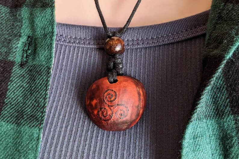 May include: A round, reddish-brown pendant necklace with a black cord. The pendant features a triple spiral design in black. The necklace is worn over a gray ribbed shirt and a green and black plaid shirt.