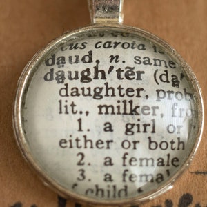 Puede incluir: Un colgante de plata con un c&iacute;rculo de vidrio que contiene una definici&oacute;n del diccionario de la palabra "hija". La definici&oacute;n dice: "hija (dɔːtə) hija, probablemente lit., orde&ntilde;adora, de 1. una ni&ntilde;a o una u otra o ambas 2. una mujer 3. una ni&ntilde;a"