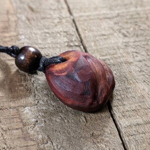 May include: A handcrafted wooden pendant necklace. The pendant is a dark reddish-brown color with a carved design and a hole at the top. It hangs from a black cord with a wooden bead. The necklace is displayed on a wooden surface.