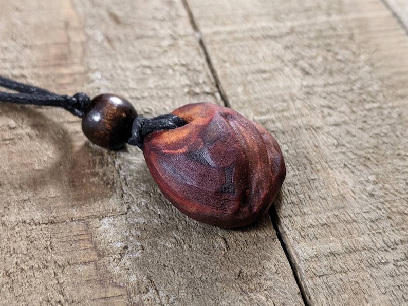 May include: A handcrafted wooden pendant necklace. The pendant is a dark reddish-brown color with a carved design and a hole at the top. It hangs from a black cord with a wooden bead. The necklace is displayed on a wooden surface.