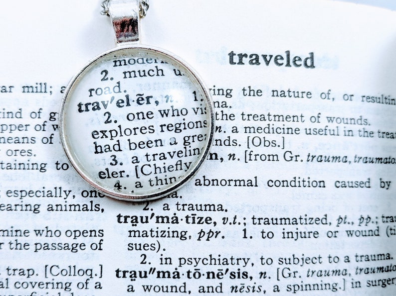 May include: A silver necklace with a round pendant featuring the word "traveler" defined in a vintage dictionary. The definition includes the words "one who explores regions" and "a traveling elder".