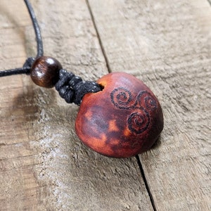 May include: A handmade pendant necklace featuring a reddish-brown, oval-shaped pendant with a carved spiral design. The pendant is strung on a black cord with a wooden bead accent. The necklace is displayed on a wooden surface.