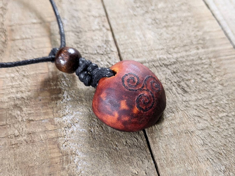 May include: A handmade pendant necklace featuring a reddish-brown, oval-shaped pendant with a carved spiral design. The pendant is strung on a black cord with a wooden bead accent. The necklace is displayed on a wooden surface.