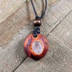 Pu&ograve; includere: Collana con pendente in legno fatto a mano con cristallo di quarzo naturale. Il pendente &egrave; di colore marrone-rossastro con una texture intagliata e un centro di cristallo trasparente. La collana ha un cordino nero e una perla di legno.