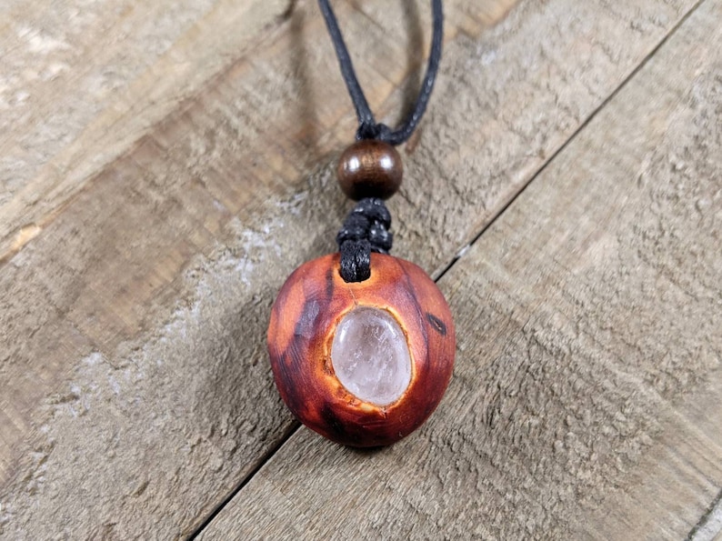 Pu&ograve; includere: Collana con pendente in legno fatto a mano con cristallo di quarzo naturale. Il pendente &egrave; di colore marrone-rossastro con una texture intagliata e un centro di cristallo trasparente. La collana ha un cordino nero e una perla di legno.
