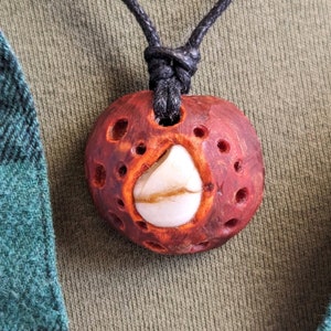 May include: A red wooden pendant with a white stone inlay and a black cord. The pendant has a carved design with multiple holes.