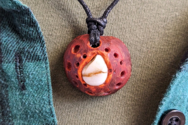 May include: A red wooden pendant with a white stone inlay and a black cord. The pendant has a carved design with multiple holes.